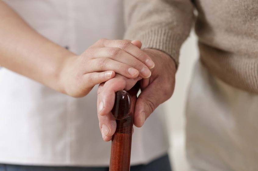 An older person holds a walking stick with a younger person carefully placing their hand on theirs